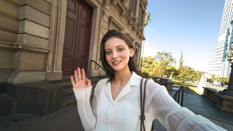 A young woman with long brown hair, wearing a white blouse, smiles and waves at the camera while standing outside a historic building on a sunny day. Trees and modern buildings are visible in the background.