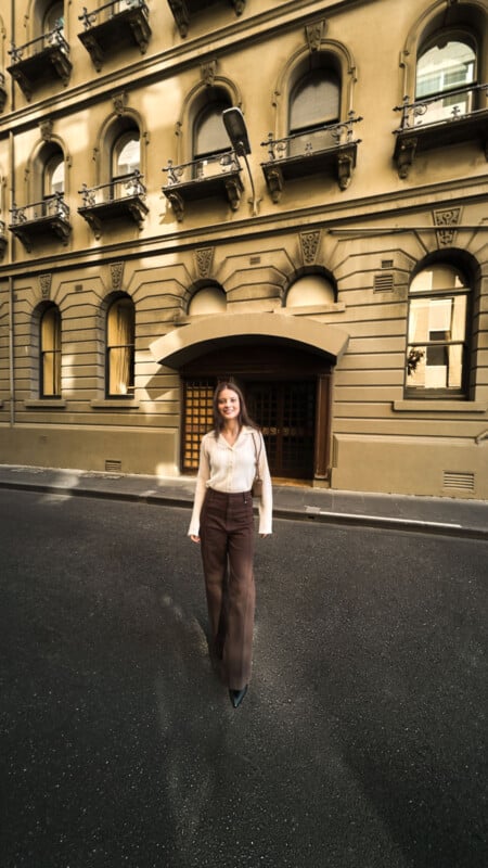 A woman wearing a light blouse and brown pants stands smiling on an empty street in front of an ornate, historic building with arched windows and balconies.
