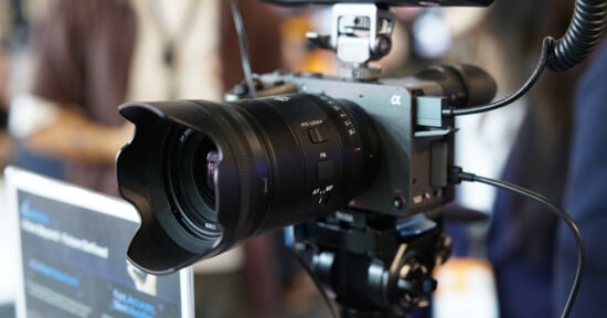Close-up of a professional digital camera with a large lens and lens hood mounted on a tripod, surrounded by cables, set up indoors with a blurred background.