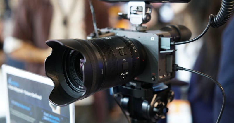 Close-up of a professional digital camera with a large lens and lens hood mounted on a tripod, surrounded by cables, set up indoors with a blurred background.