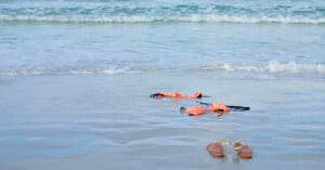 A pair of sandals and a floral bikini lie on the wet sand near the shoreline, with gentle ocean waves approaching in the background.