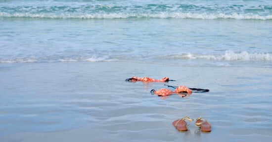 A pair of sandals and a floral bikini lie on the wet sand near the shoreline, with gentle ocean waves approaching in the background.