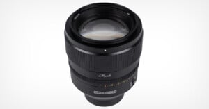 A close-up view of a black camera lens with textured focus rings, engraved markings, and the brand name “Monolith” visible on the barrel against a plain white background.
