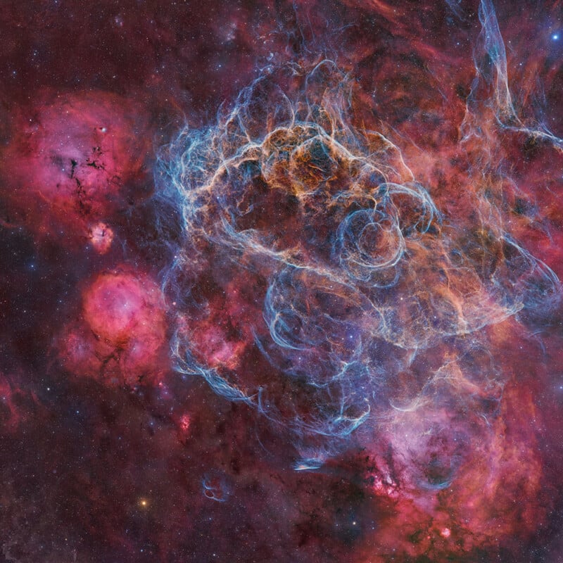 Colorful nebula in deep space, featuring bright pink and red clouds with intricate, wispy blue filaments forming swirling patterns against a dark star-filled background.
