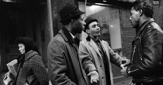 Two men stand talking on a city sidewalk while a third man in a leather jacket listens; a woman in a coat and headscarf walks past carrying newspapers. The scene is set in front of urban storefronts.