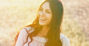 A woman with long dark hair wearing a white blouse smiles brightly in soft, golden sunlight, with an out-of-focus natural background.