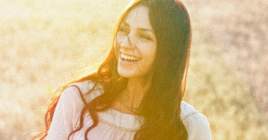 A woman with long dark hair wearing a white blouse smiles brightly in soft, golden sunlight, with an out-of-focus natural background.