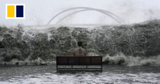 A person sits alone on a bench facing a massive, crashing wave with a bridge in the background, under a gray sky. A blue and yellow rectangular logo appears in the upper left corner.