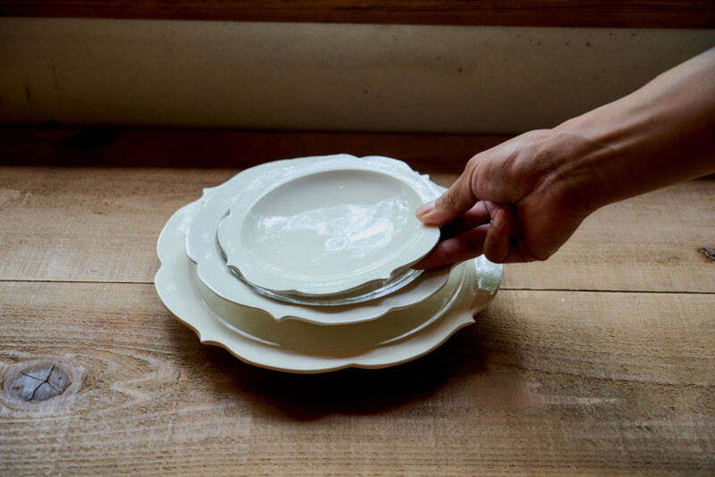 A hand is stacking white ceramic plates of various sizes on a wooden table. The plates are neatly nested together, and the background shows a wooden surface and a windowsill.