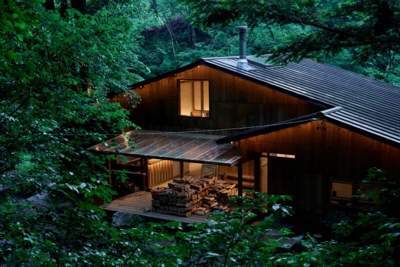 A wooden cabin with warm lights glows among lush green trees; stacks of firewood are neatly arranged under a covered porch, creating a cozy, secluded atmosphere in the forest.