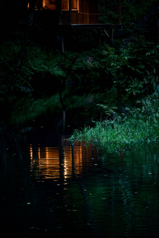 A warm light from a house reflects on the dark, calm surface of a river surrounded by dense greenery at night.