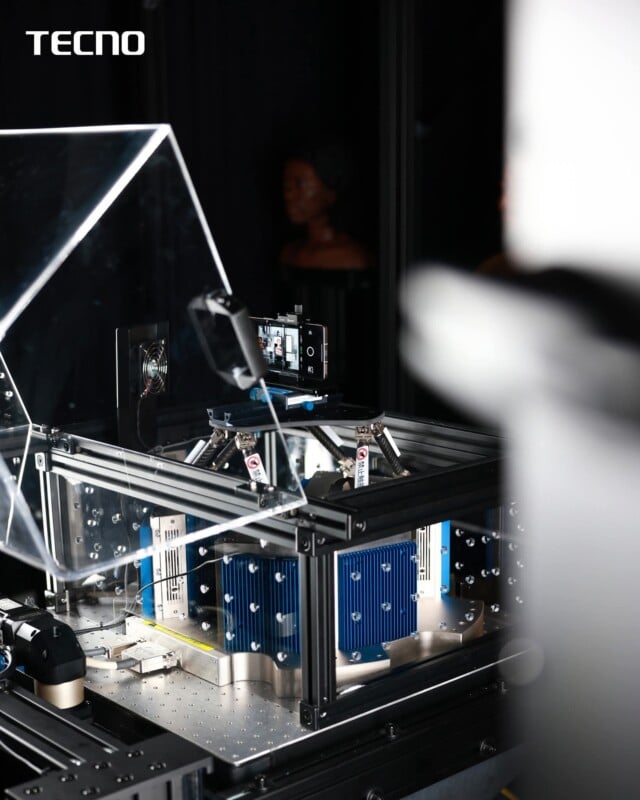 Se instala un teléfono inteligente en una plataforma de prueba transparente de alta tecnología con un radiador azul marcado con una etiqueta "tecno" En la esquina superior izquierda. La configuración parece estar en un entorno de laboratorio.