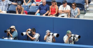 Four photographers with large cameras aim their lenses at an event below, while a row of spectators sits in the stadium seats above, watching intently.