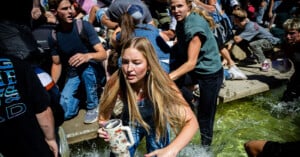 A group of young people scramble through shallow water, looking distressed and hurried. A girl in the foreground holds a water bottle, her long hair wet, as others crowd around her in a chaotic outdoor scene.