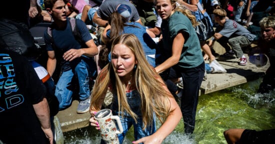 A group of young people scramble through shallow water, looking distressed and hurried. A girl in the foreground holds a water bottle, her long hair wet, as others crowd around her in a chaotic outdoor scene.