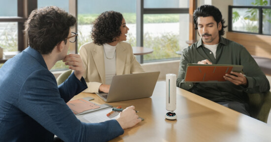 Three people sit around a table in an office, having a discussion. One person writes on a notepad, another listens with a laptop open, and a third observes. A small, white conference microphone sits on the table.