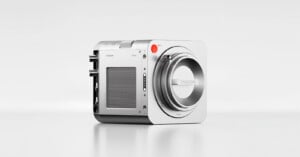 A modern, compact digital camera with a sleek, square design, silver body, and prominent lens mount. The camera features a red record button and minimalistic controls against a plain white background.