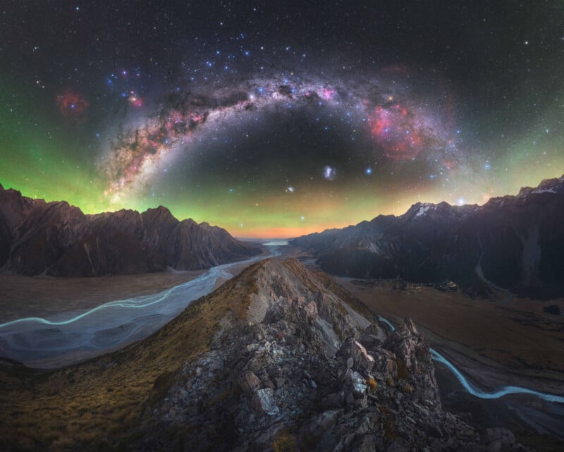 A rocky mountain ridge stretches toward a winding river under a vibrant night sky, featuring the Milky Way arching over jagged peaks and colorful auroras glowing on the horizon.
