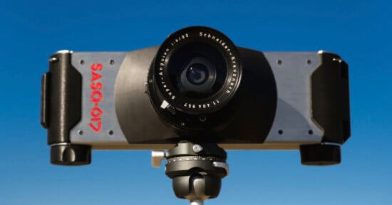 A close-up of a SASQ-617 panoramic film camera mounted on a tripod, with a wide-angle lens facing forward against a clear blue sky.