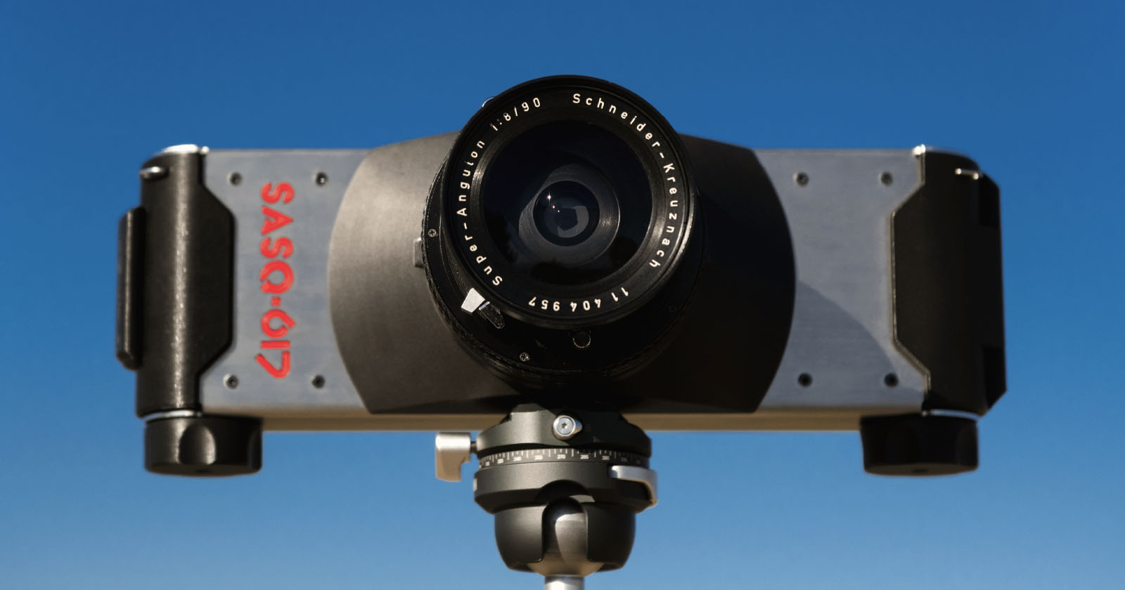 A close-up of a SASQ-617 panoramic film camera mounted on a tripod, with a wide-angle lens facing forward against a clear blue sky.