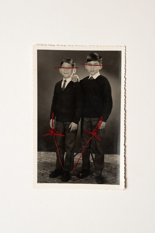 A black-and-white vintage photo of two boys in sweaters and ties. Red thread is stitched across their eyes and hands, with lines extending down to their feet, giving the photo an altered, surreal effect.