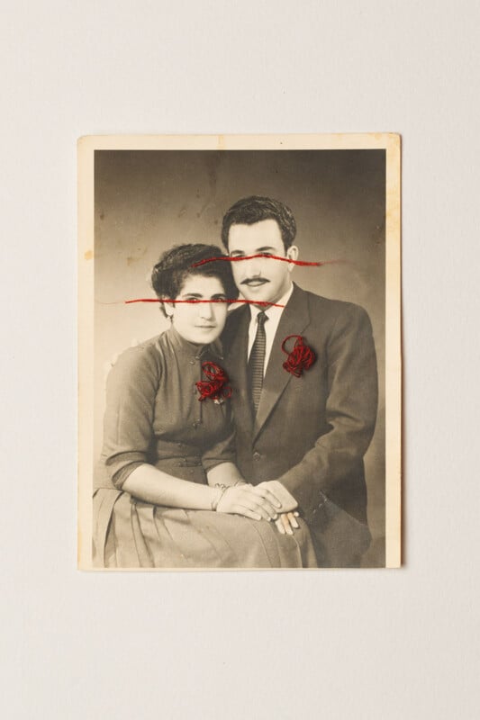 A vintage black-and-white portrait of a seated couple with red embroidery over their eyes and hearts. The photograph has slightly worn edges and is set against a plain light background.