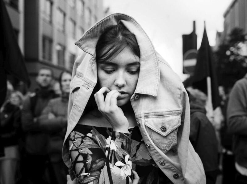 A young woman with a thoughtful expression stands in a crowd, draping a denim jacket over her head. She wears a floral-patterned top, and the scene is set on a city street in black and white.