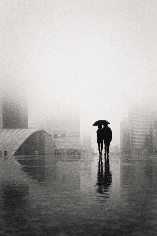 Two people stand close together under an umbrella on a wet, reflective surface in a cityscape shrouded in thick fog, with tall buildings barely visible in the background.