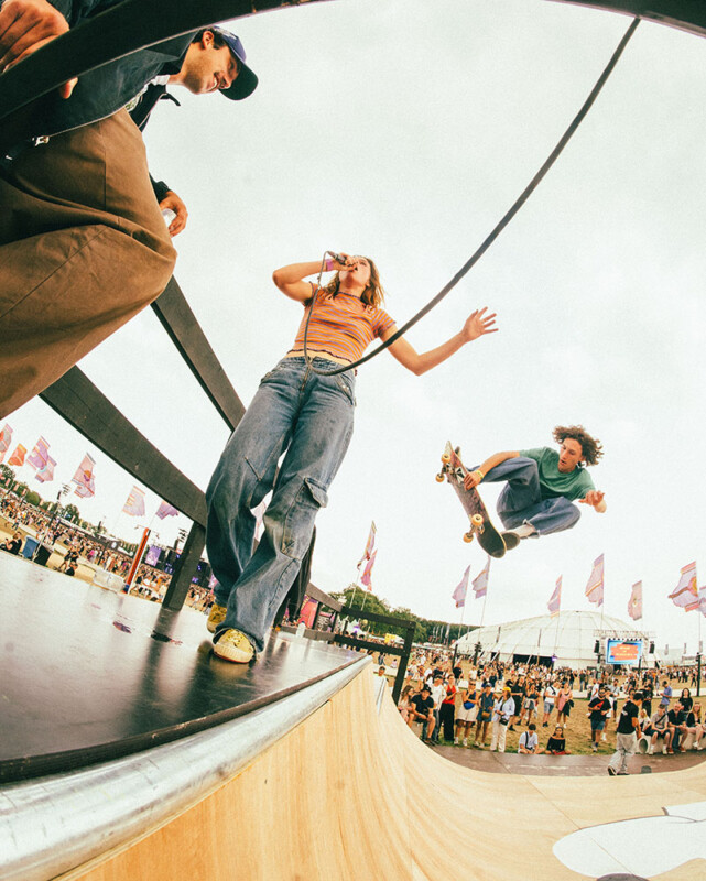 El patinador muestra la habilidad de saltar de la rampa cuando la mujer canta en el micrófono y el hombre se sienta cerca. Las multitudes y los pancartas del festival llenan el fondo nublado.
