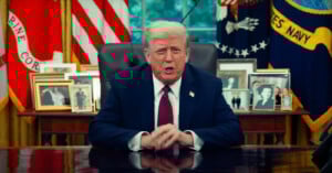 A man in a dark suit and red tie sits at a desk in an office, with framed photos, American flags, and military banners in the background.