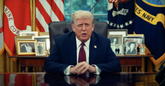 A man in a dark suit and red tie sits at a desk in an office, with framed photos, American flags, and military banners in the background.