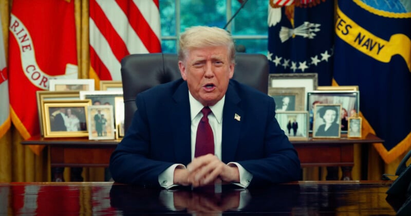 A man in a dark suit and red tie sits at a desk in an office, with framed photos, American flags, and military banners in the background.
