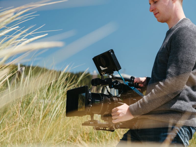 A person wearing a gray sweater operates a professional video camera outdoors, surrounded by tall grass under a clear blue sky.