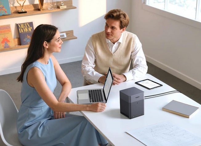 Dos personas sentadas en la mesa blanca en la oficina moderna hablando. La mujer usa la computadora portátil, el hombre tiene una tableta. Tabla con libros, cuaderno y equipo, estantes y ventanas en el fondo.