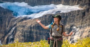A park ranger with braided hair gestures while speaking, standing outdoors with a rocky mountain and glacier in the background. She wears a green uniform, cap, and backpack.