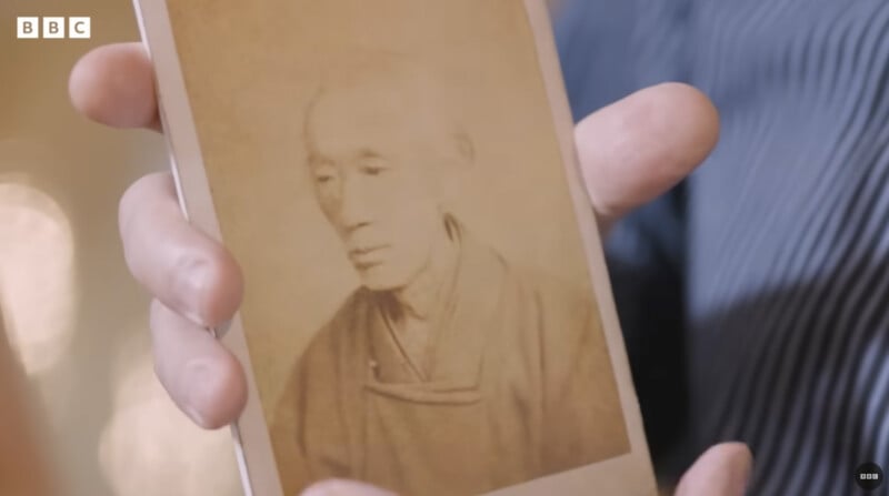A hand holds a sepia-toned photograph of an elderly person wearing traditional clothing. The background is blurred, and the BBC logo appears in the upper left corner.