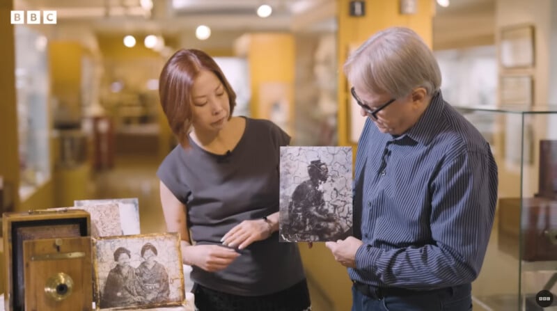 Two people stand in a museum, examining old black-and-white photographs. One person holds a photo, while the other looks on. Vintage cameras and more photos are displayed on the table beside them. BBC logo is visible in the corner.