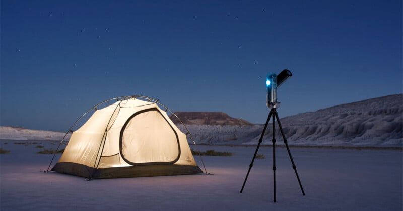 A glowing tent stands on a flat, sandy landscape under a clear night sky with stars, next to a telescope on a tripod pointing upward; distant hills are visible in the background.