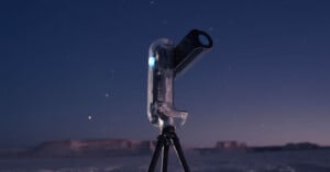 A modern, transparent telescope on a tripod stands outdoors at dusk, with a clear sky and distant mesas in the background. Stars are visible in the sky, and the scene has a calm, twilight atmosphere.