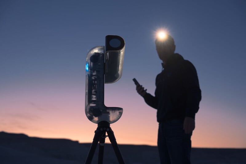 A person wearing a headlamp stands outdoors at dusk, holding a device next to a large, transparent scientific instrument mounted on a tripod. The sky is a gradient of purple and orange as the sun sets.
