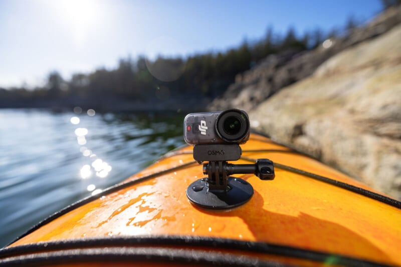 A small action camera mounted on the front of an orange kayak, floating on a calm lake with rocky shoreline and trees in the background, under a clear blue sky.