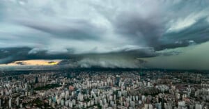 A dramatic, dense storm cloud hovers over a sprawling city skyline at sunset, with tall buildings below and dark, turbulent clouds dominating the sky.