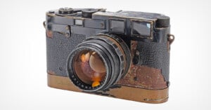 A vintage Leica camera with a worn, black and brass body and a textured grip. The lens is prominent, and the exterior shows signs of heavy use and age, revealing patches of metal beneath the original finish.