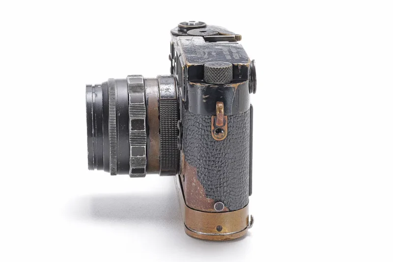 A vintage, weathered camera with a textured black body and metallic accents is shown from the side on a white background. The lens is extended, and signs of wear are visible on the body.