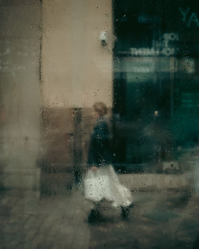 A blurry figure of a person in a white skirt walking outside, seen through a rain-streaked window with water droplets. The background features a city street and building facade.