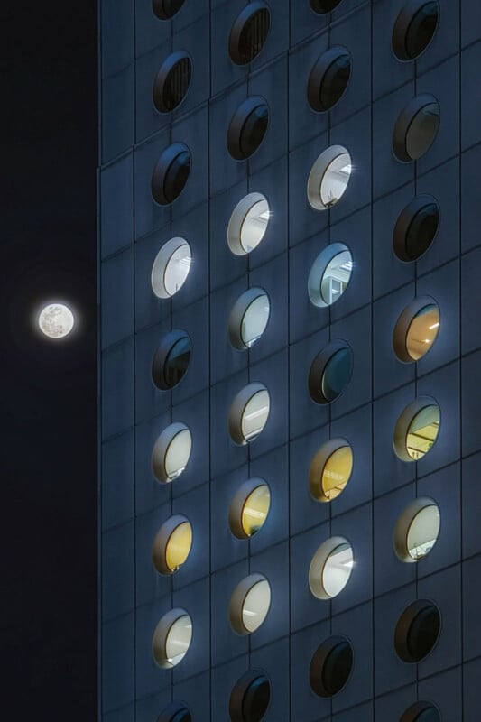 A modern building facade with round windows, some lit from within, is shown at night. The full moon is visible in the dark sky to the left of the structure.