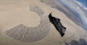 A wingsuit flyer soars above an aerial view of Black Rock City, a temporary circular city in the Nevada desert, with camps and streets visible below and barren desert surrounding the area.