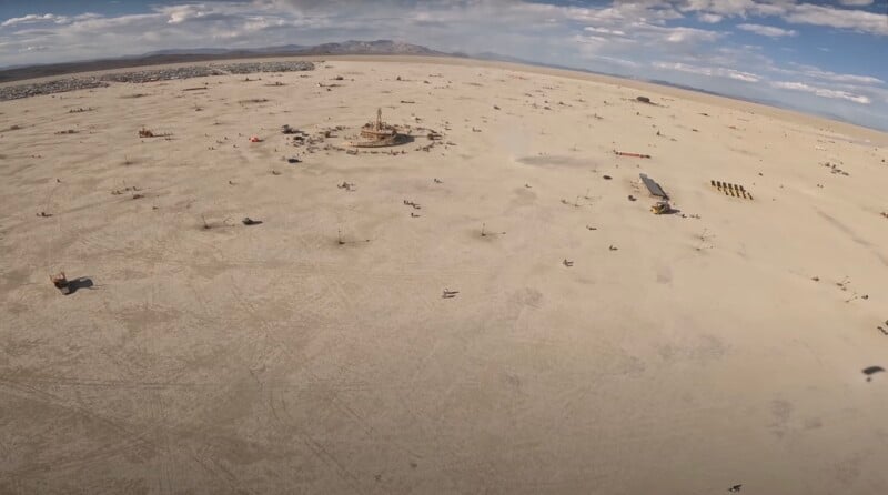 A wide aerial view of a sandy desert plain with scattered people, vehicles, and temporary structures, under a partly cloudy sky with distant mountains on the horizon.