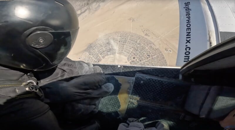 A skydiver in black gear prepares to jump from a plane, with a bird’s-eye view of a circular desert settlement visible far below. The plane’s doorway and “SkydivePhoenix.com” text are also visible.