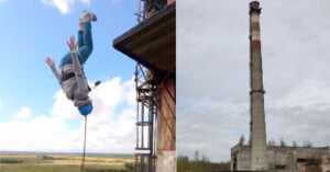 A person wearing a helmet and harness jumps off the side of a tall industrial building, upside down in mid-air; beside is a tall, old smokestack rising above an abandoned structure.